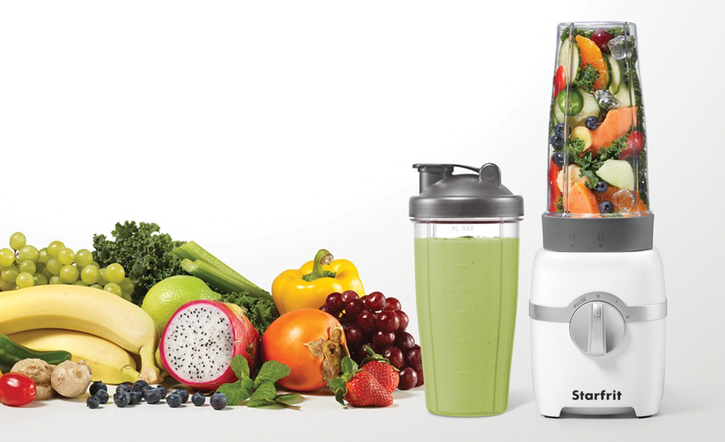 A small blender sitting on a countertop beside a cornucopia of fruits and vegetables.