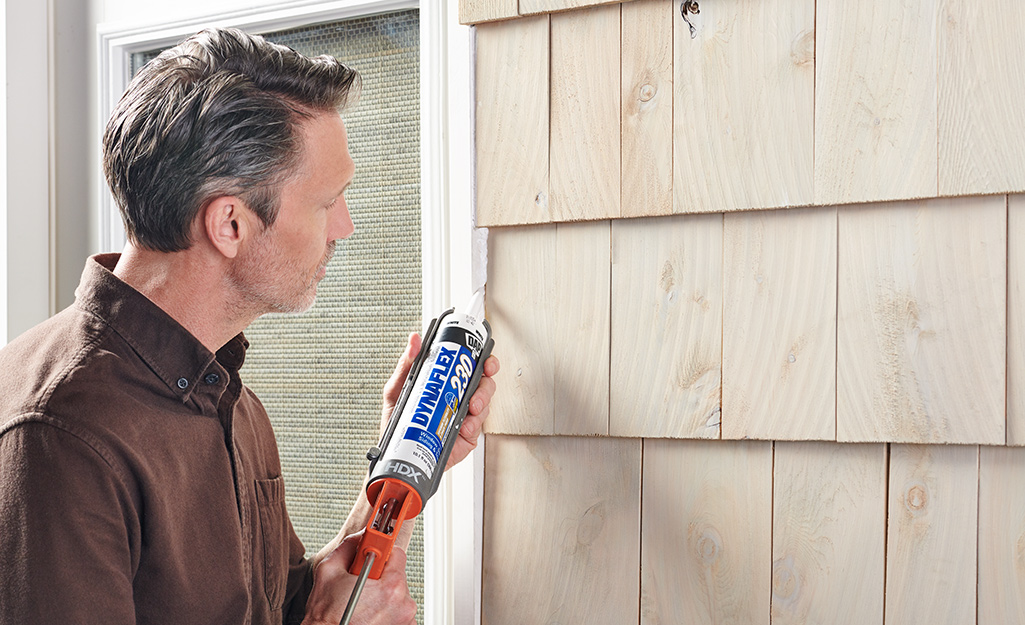 A person using caulk to seal between siding and a window frame.