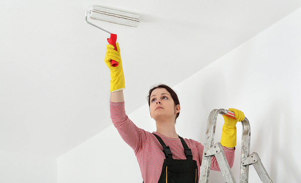 A woman on a ladder uses a paint roller to paint a ceiling.