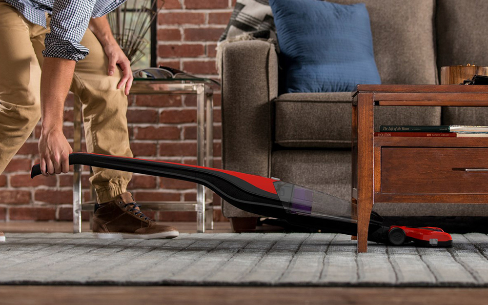 A person pushing a cordless vacuum on a rug under a table