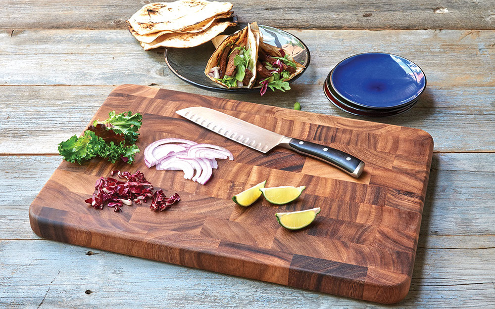 An end-grain wood cutting board topped with a knife and cut produce.