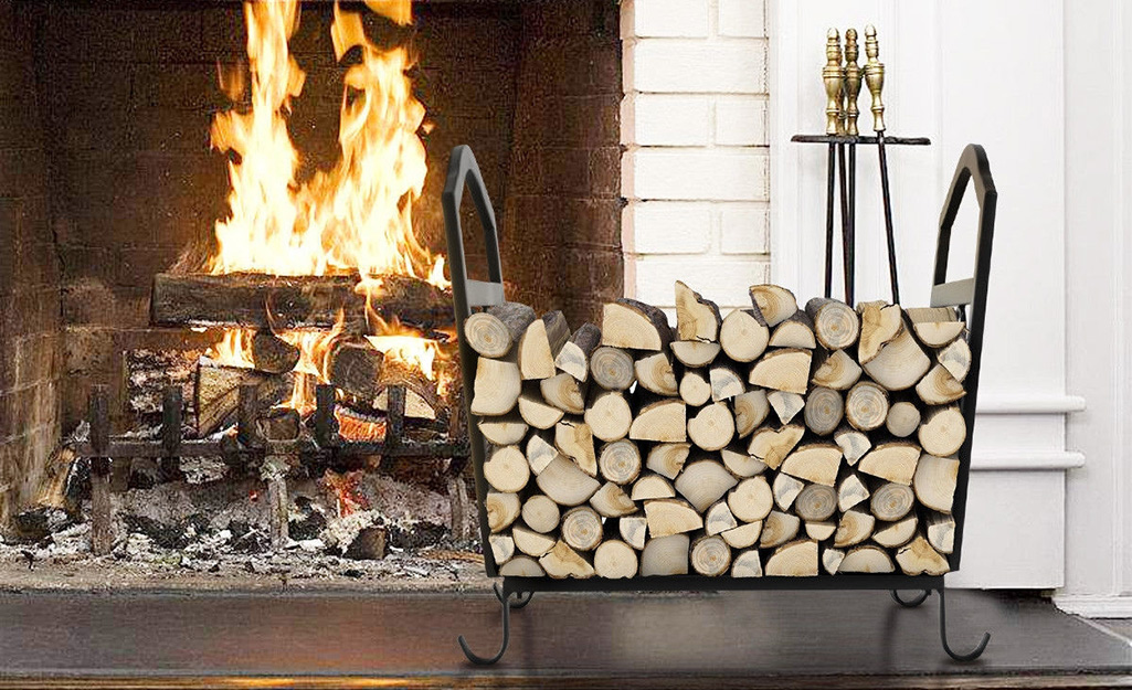 A stack of cut logs sits in a wood rack next to a roaring fireplace.