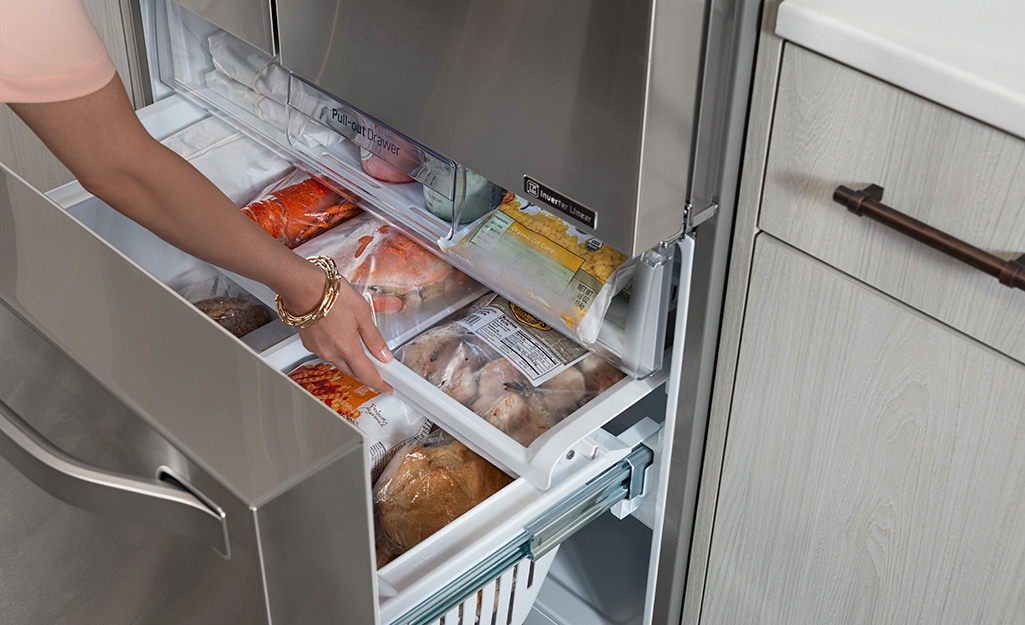 A person reaches into an open sliding drawer in the bottom half of an upright freezer.