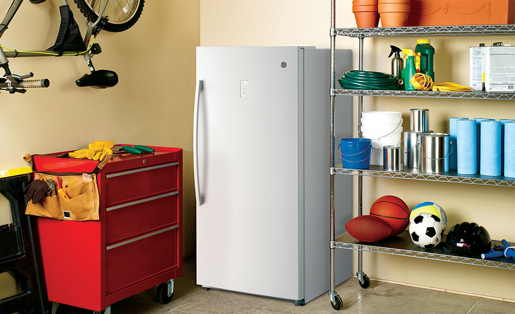 An upright freezer stands in the corner of a garage next to a red tool chest and a wire shelving unit.
