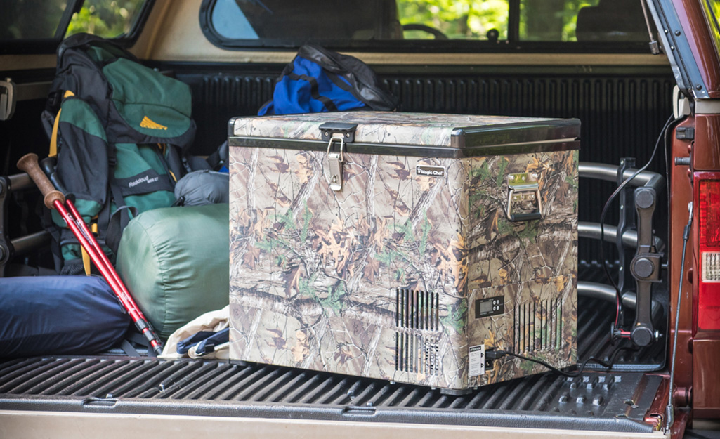 A portable freezer with a camouflage design sits next to camping gear in the bed of a pickup truck.