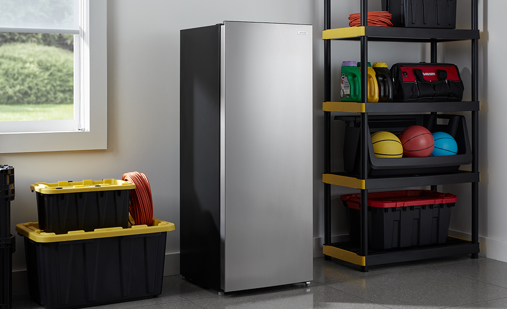 A stainless steel freezer stands in a garage next to plastic storage boxes and a black-and-yellow shelving unit.