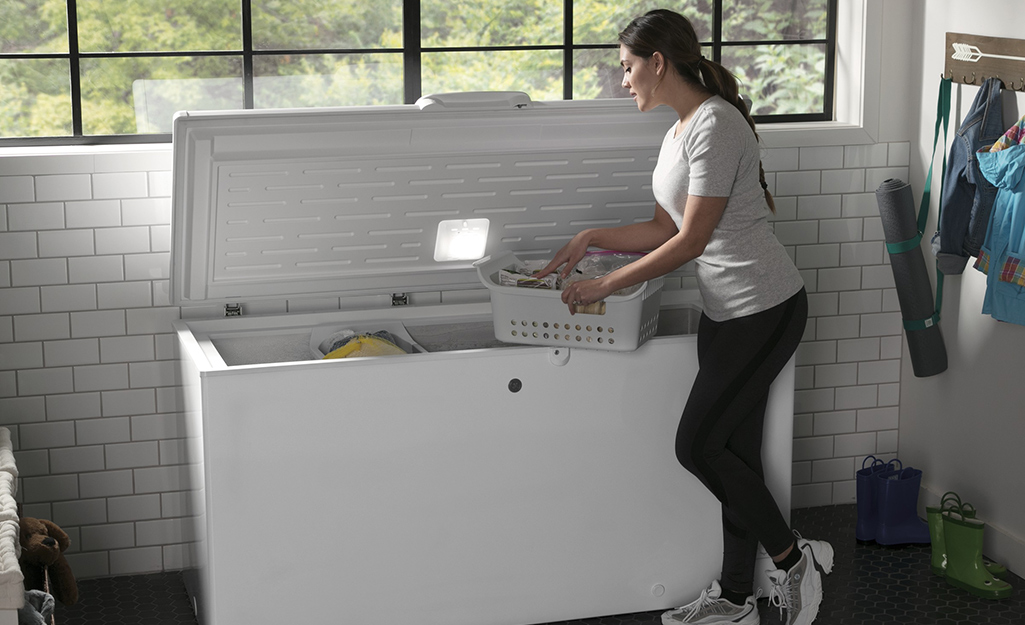 A woman looks at the contents of a white plastic bin while holding it on the edge of an open chest freezer.