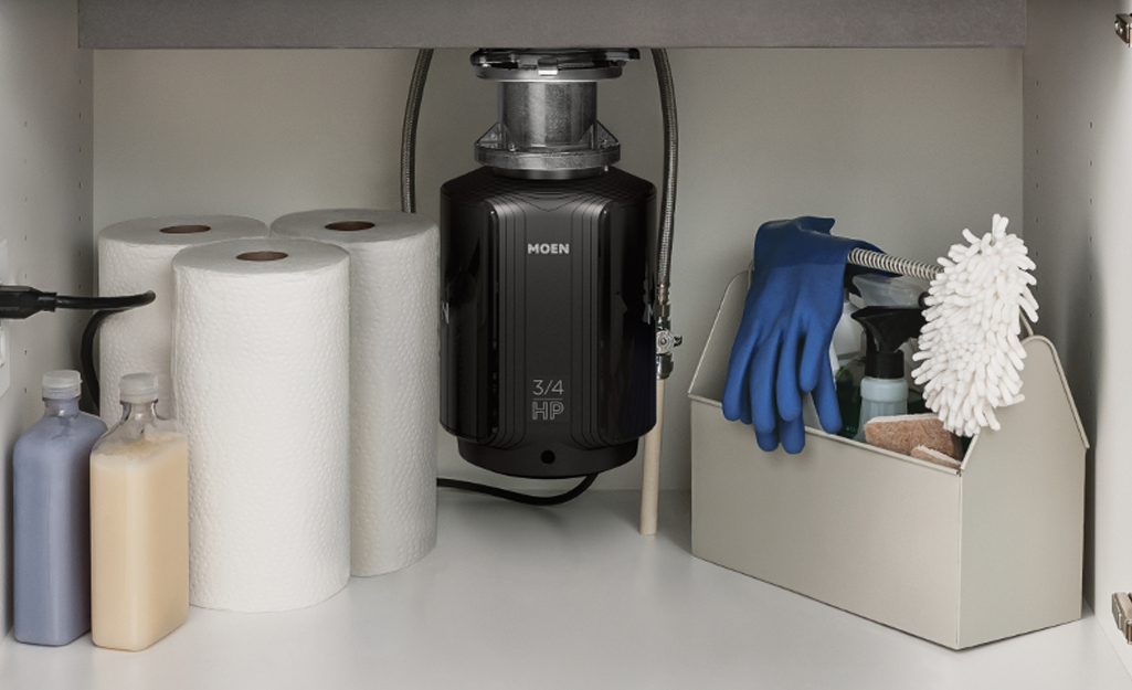 A batch feed disposal installed underneath a kitchen sink beside some paper towels and cleaning supplies.