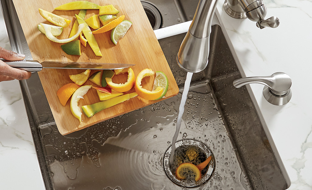 Someone using a knife to scrap citrus peelings off a cutting board into the opening of a garbage disposal while water is running.