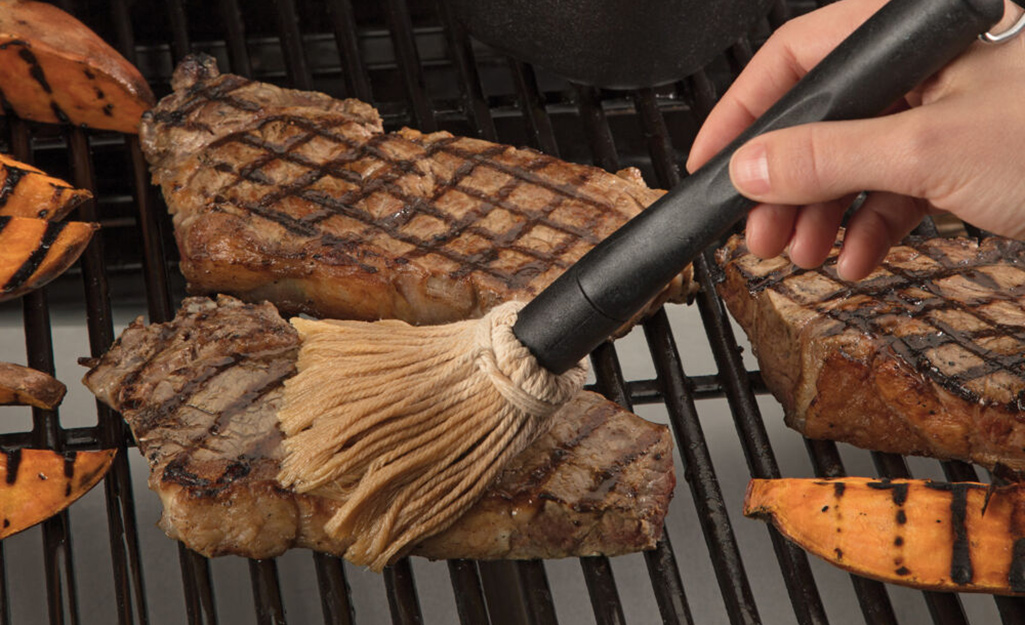 A person uses a basting brush on a steak cooking on a grill.