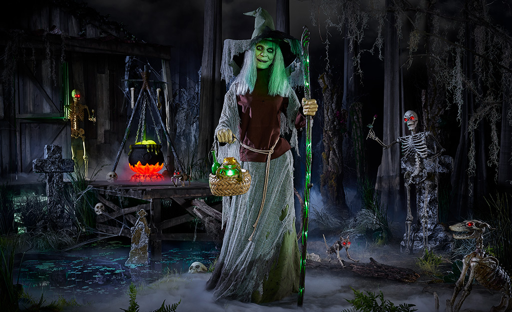 An animatronic witch with eyes that move holds a lit staff and stands in the center of a scary scene in a yard decorated for Halloween. 