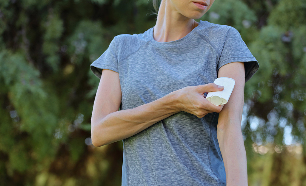 A woman wearing gray T-shirt applies roll-on insect repellent to her arm.