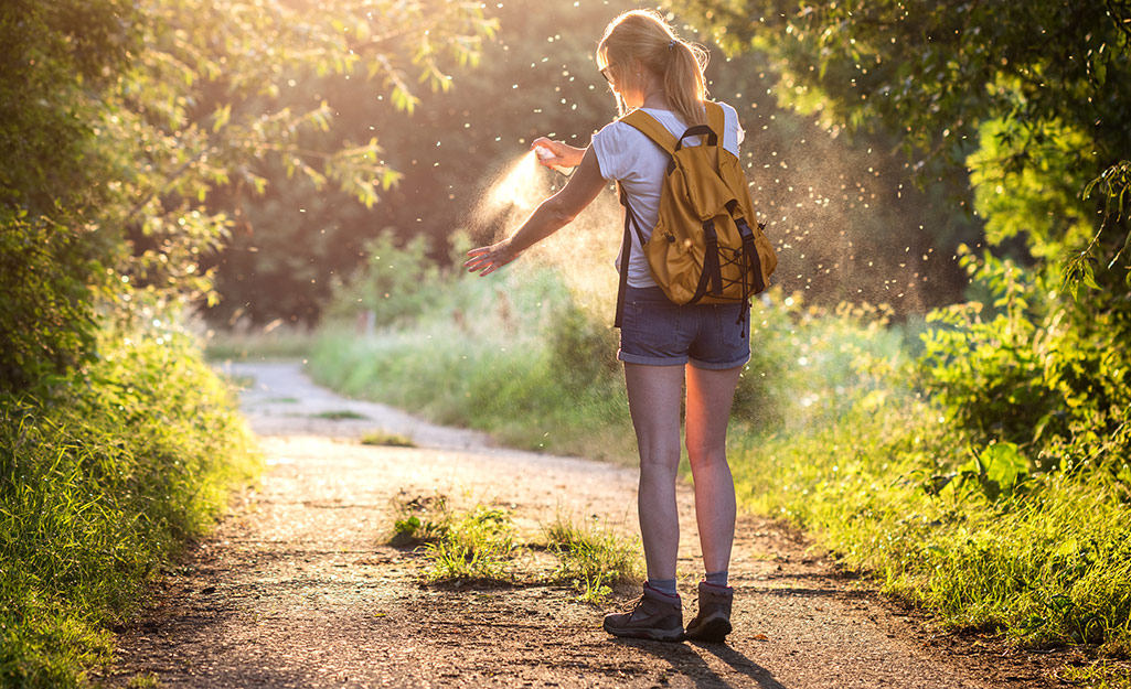 A woman with a backpack stands on a sunlit hiking trail and sprays insect repellent on her arm.