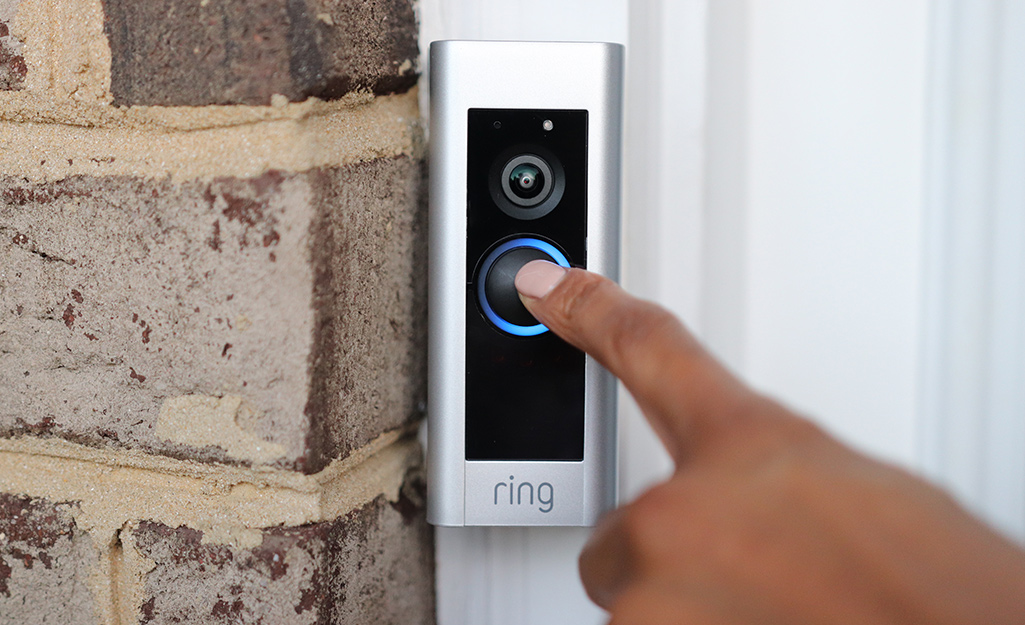 A person uses their index finger to press the button on a smart doorbell on a brick wall. 
