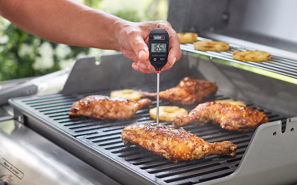 An instant-read digital thermometer measures the temperature of chicken on a grill.