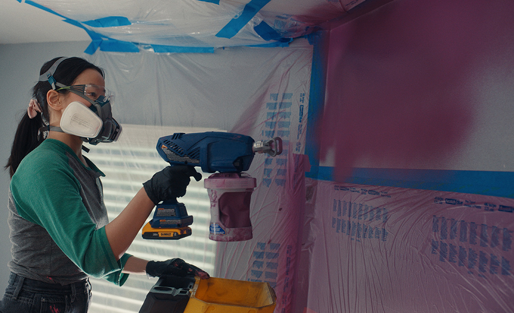 A person wearing safety googles and a respirator uses a paint sprayer to paint a wall.