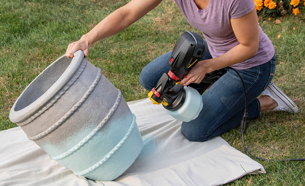 A person uses a paint sprayer to paint a large flower pot.