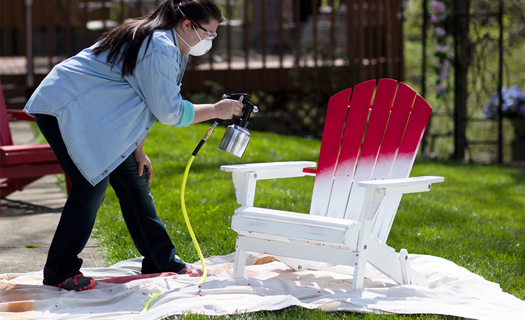 A woman uses a paint sprayer to paint an Adirondack chair.