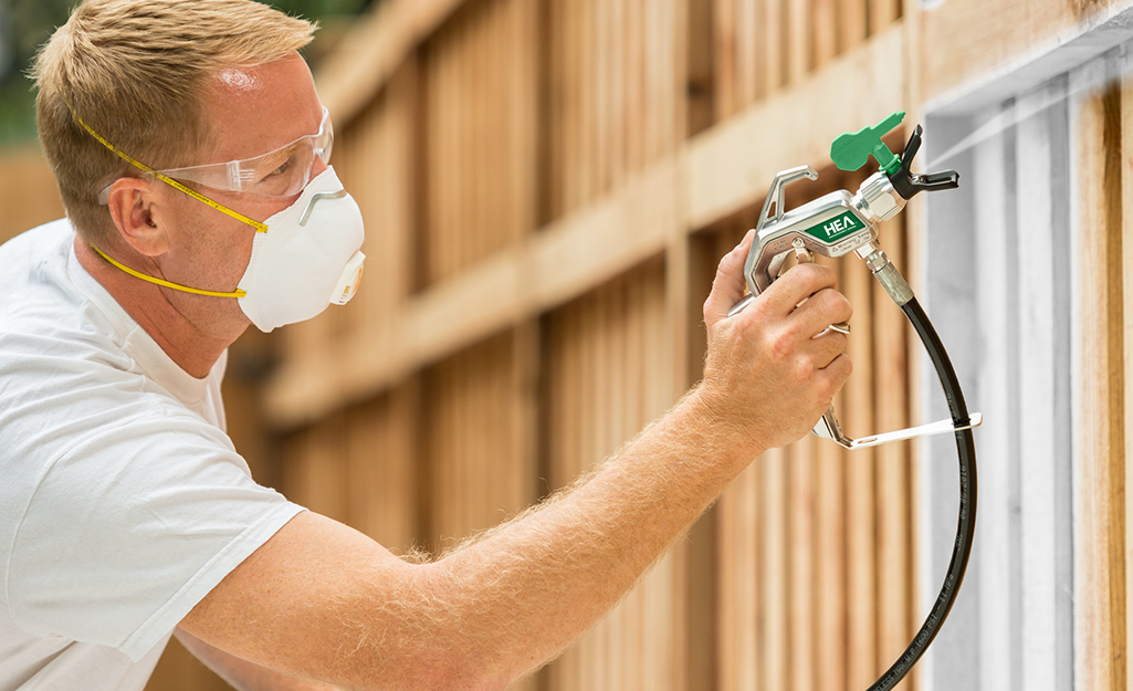 A man wearing a safety mask and protective eyewear uses a paint sprayer to paint a fence.