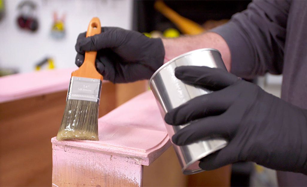 A person wearing black gloves uses a paint brush to apply paint stripper to a piece of wood furniture.