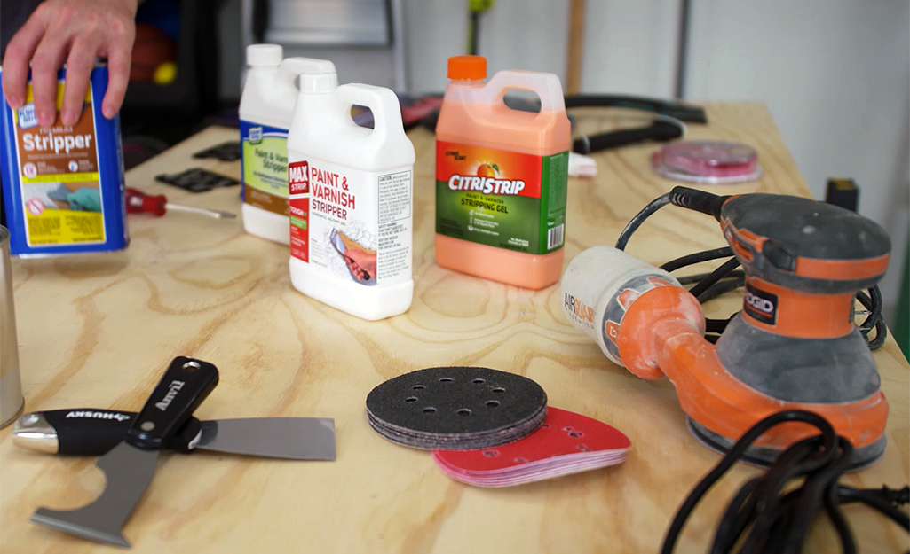A person puts a container of paint stripper down next to other solvents and tools for paint removal on a workshop table. 