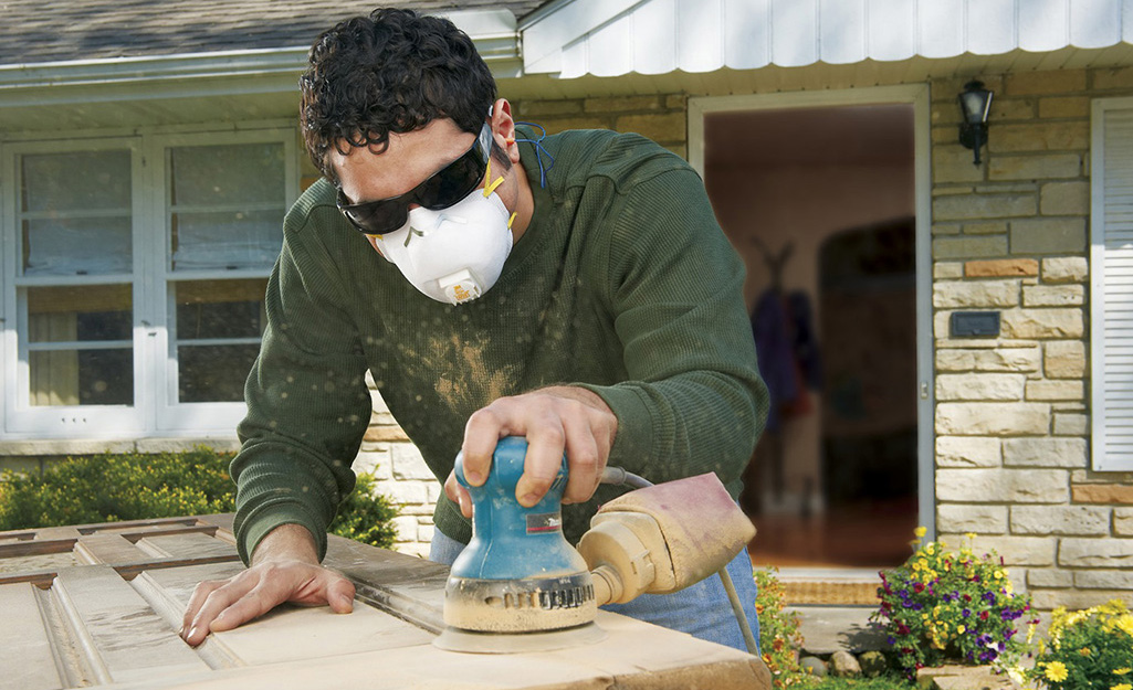 A person wearing a protective mask works outside in front of a house to sand the finish off a door. 