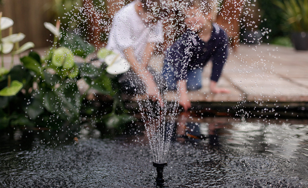 Water shoots up from a fountain in a garden pond, as an adult and a child kneel next to the pond in the background. 