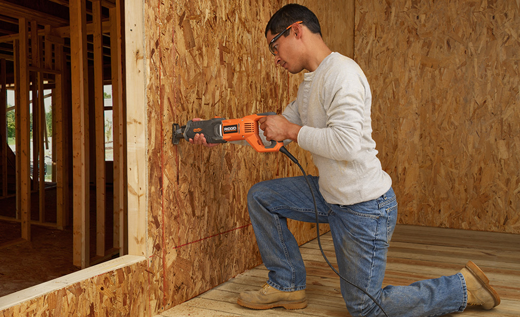 A person kneeling and using a reciprocating saw on a wall.