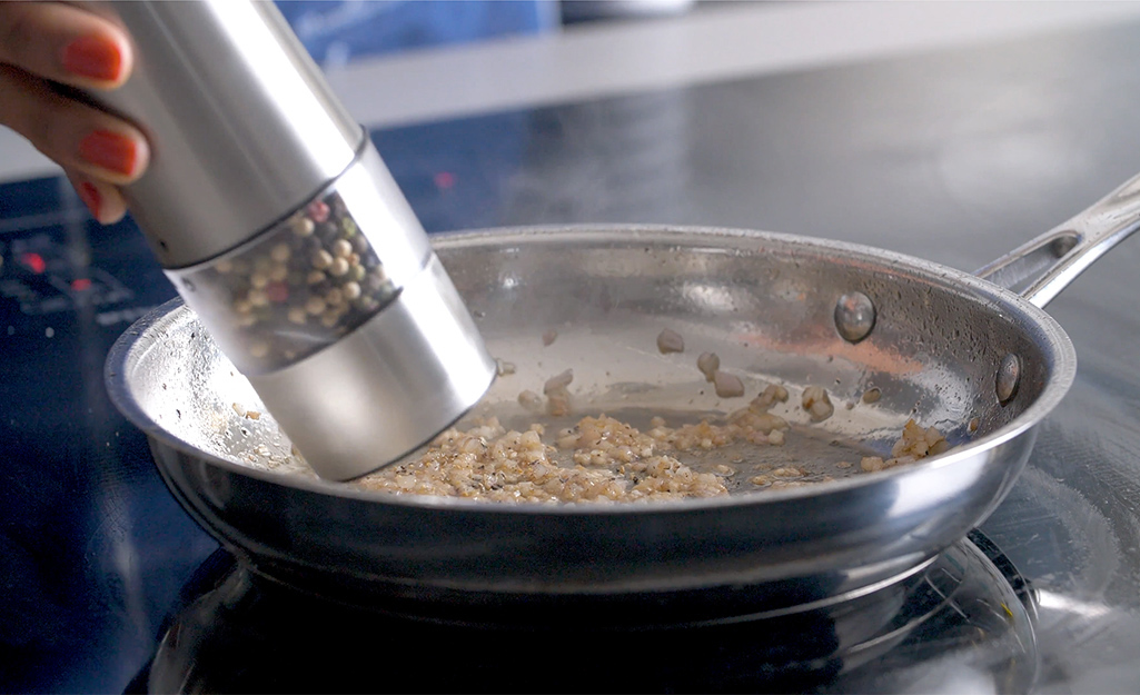 A woman uses a grinder to season a pan of sauteed garlic.