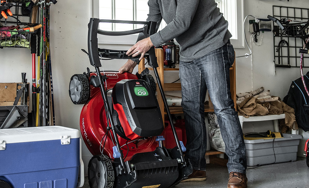 A person puts a self-propelled lawn mower into storage in a garage.
