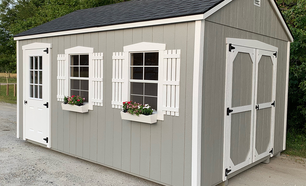 A grey shed accessorized with white faux shutters, window boxes and trim.