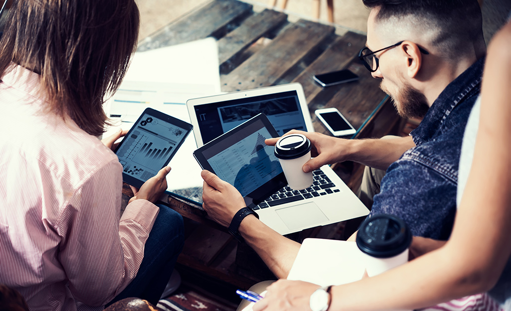 Two people monitor their smart home devices with tablets and a laptop. Two smartphones sit nearby.