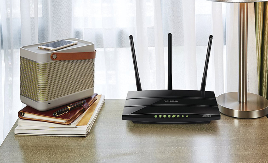 A speaker, Wi-Fi extender, smart phone, notebooks and a lamp sit on a sunny desk in front of a window.