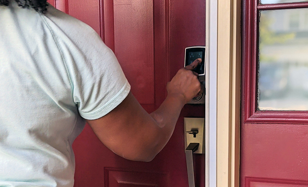 A person punches a code into the keypad of a smart lock on the red front door of a house.