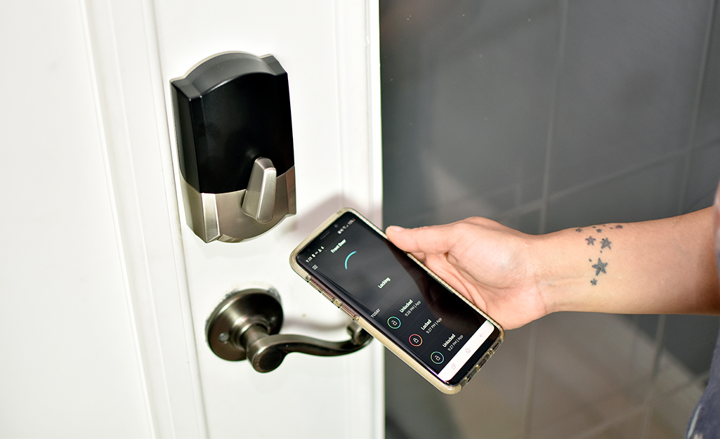 A person holds a smartphone next to the door of a house and uses an app to unlock a smart door lock.   