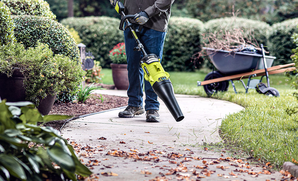 A person uses a string trimmer with a leaf blower attachment to remove leaves and debris from a paved path.