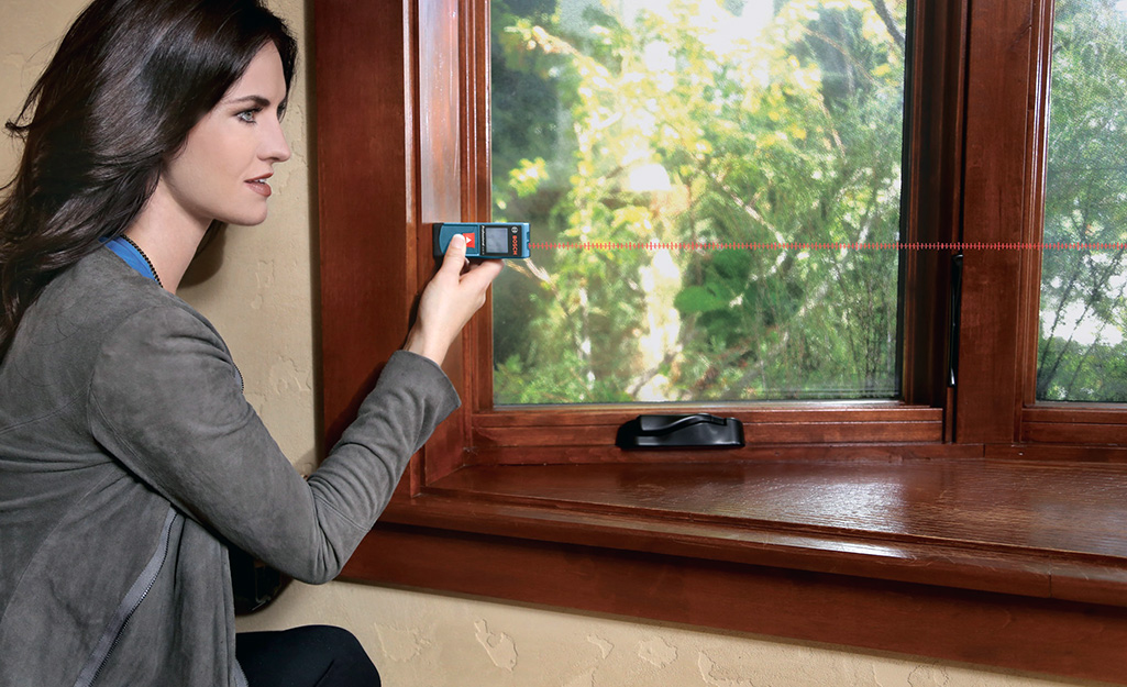 A woman using a laser tape measure on a window area.