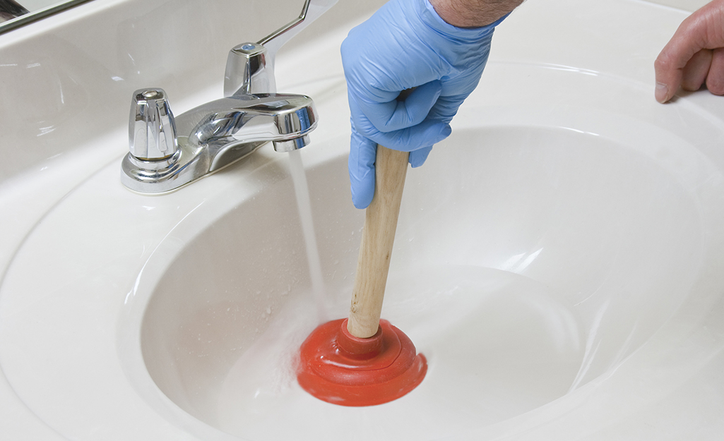 Someone wearing a blue glove and using a sink plunger under running water to unclog a sink. 
