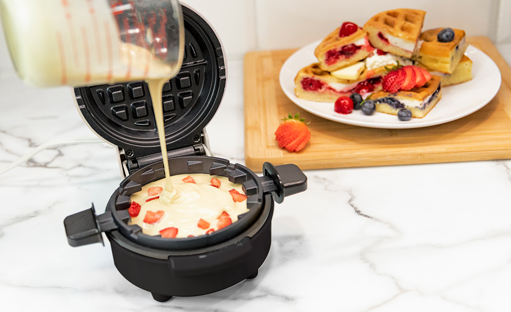 A 4-slice waffle maker with a cooked waffle inside, sitting next to a bowl of fresh fruit