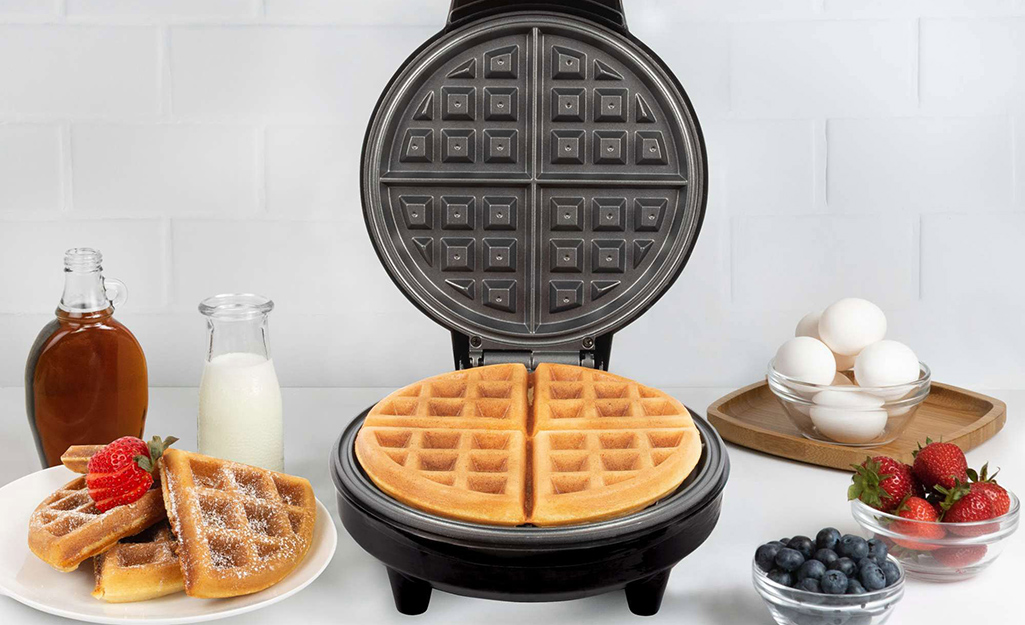 A nonstick waffle maker with a cooked waffle inside, sitting next to a cooked waffle topped with fruit and chocolate syrup
