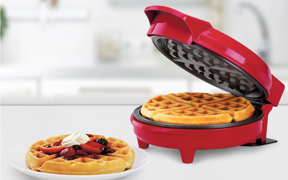 a red nonstick waffle maker with a cooked waffle inside, sitting next to a cooked waffle topped with fruit and chocolate syrup