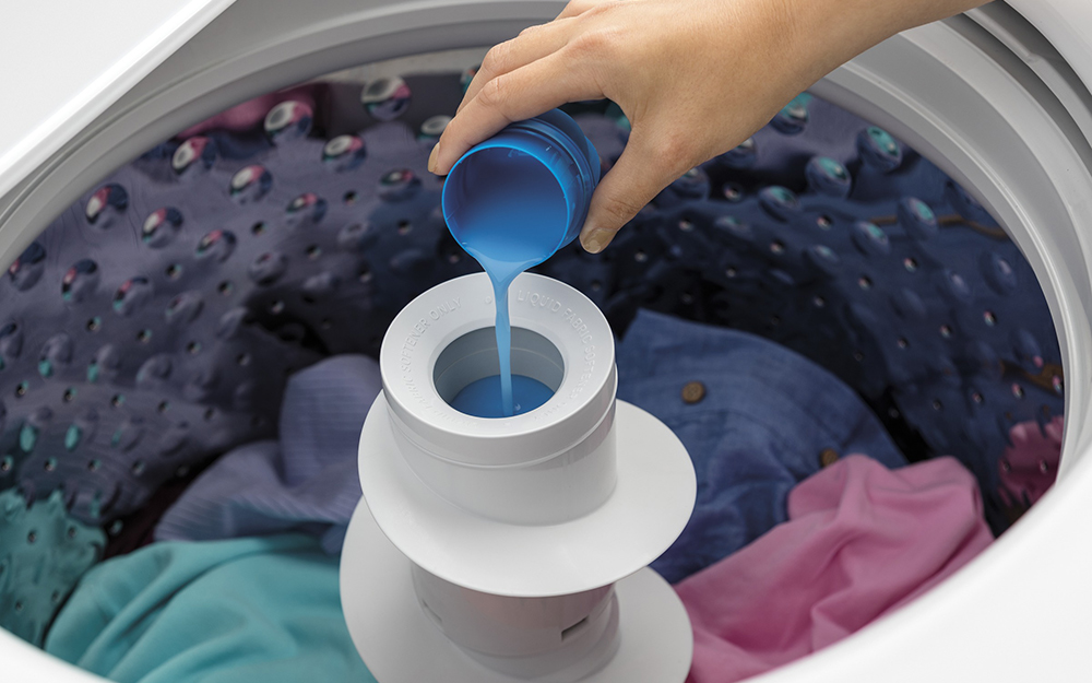 Someone adding fabric softener to a top-load washing machine filled with clothes.
