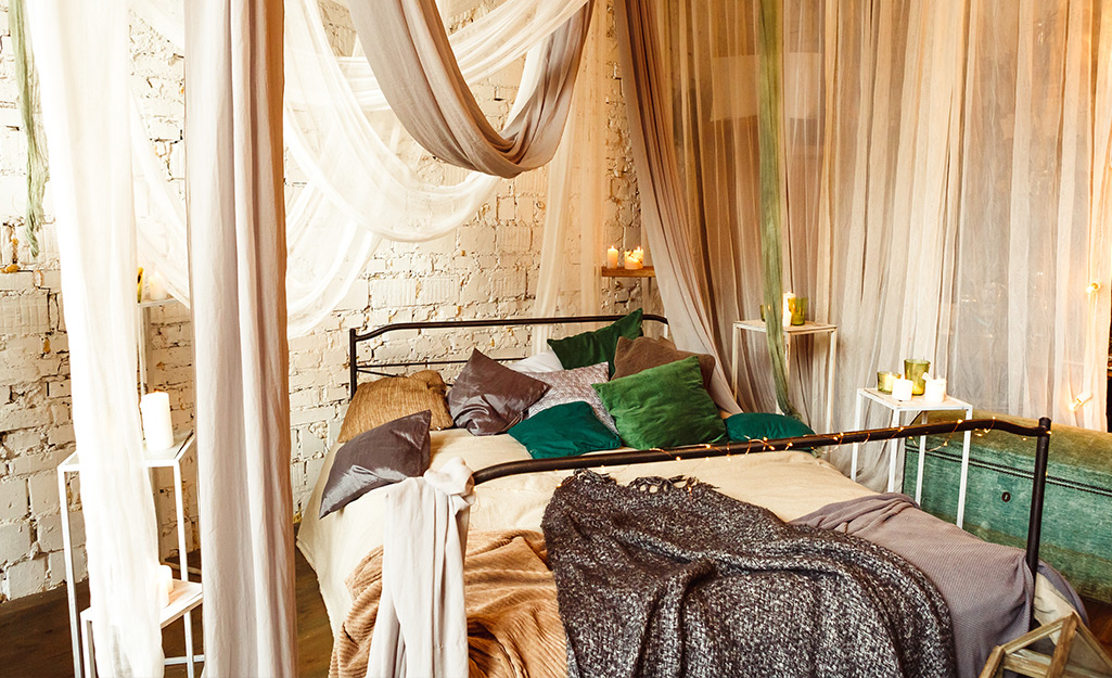 A boho styled bedroom with window curtains, fabric draped over the bed and a variety of throw pillows on the bed.