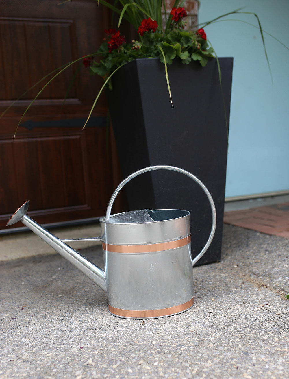 A metal watering can with copper accents sits on the ground in front of a black planter filled with flowers.