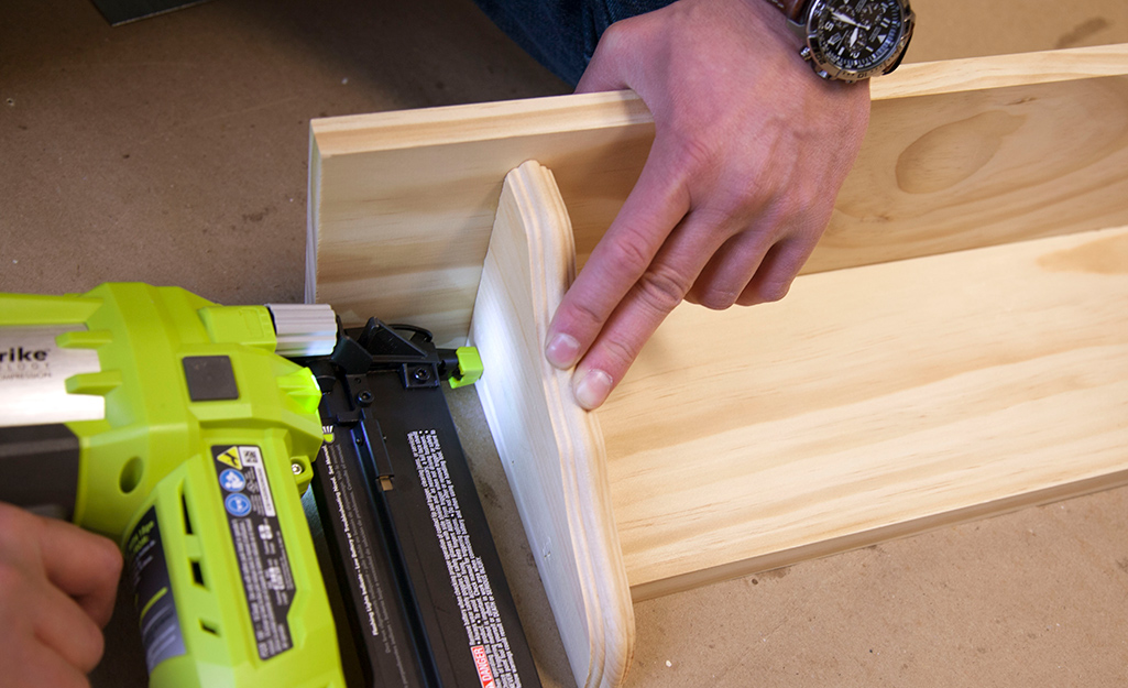 A person using a nailer with brad nails to fasten a wooden shelf together.