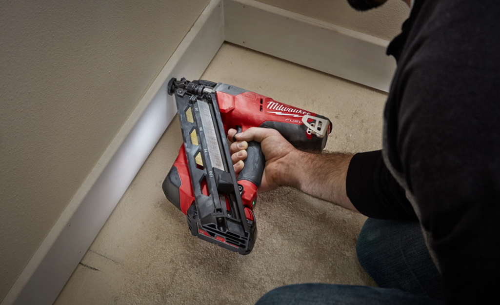 A person using a nailer to fasten baseboards to a wall.