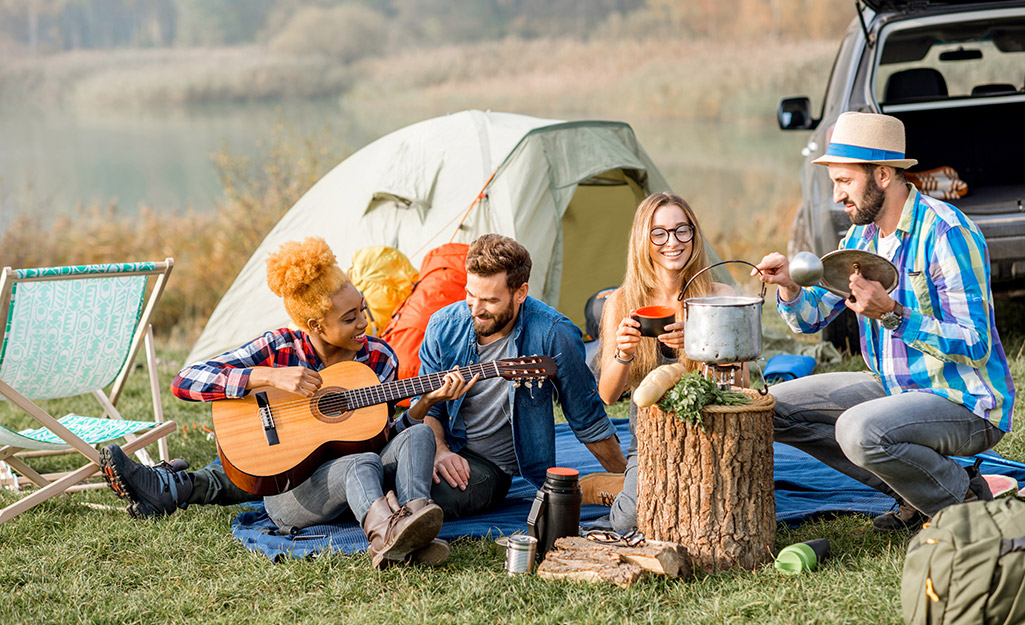 A group of campers enjoy food and music at a campsite near their tent and car.