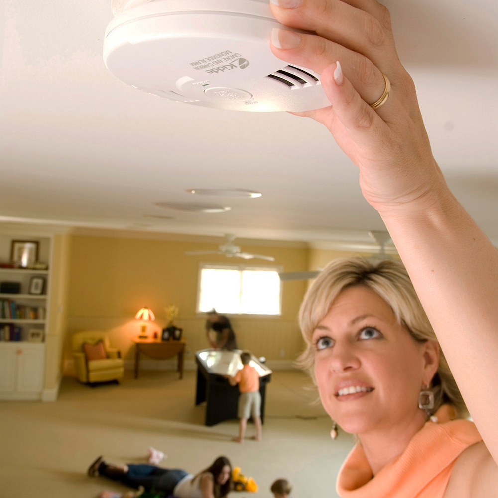 A person adjusts a carbon monoxide detector mounted on a ceiling.