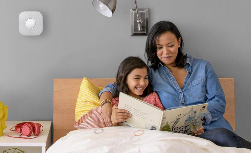 A child and adult read a book in a bed near a wall-mounted carbon monoxide detector.