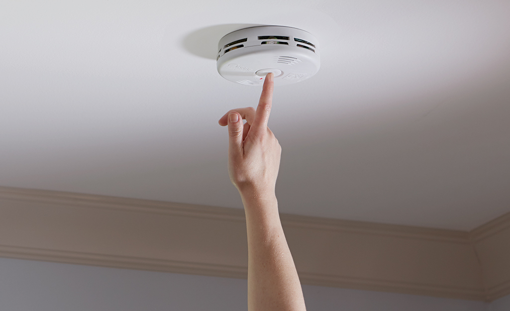 A person testing a carbon monoxide detector mounted on a ceiling.